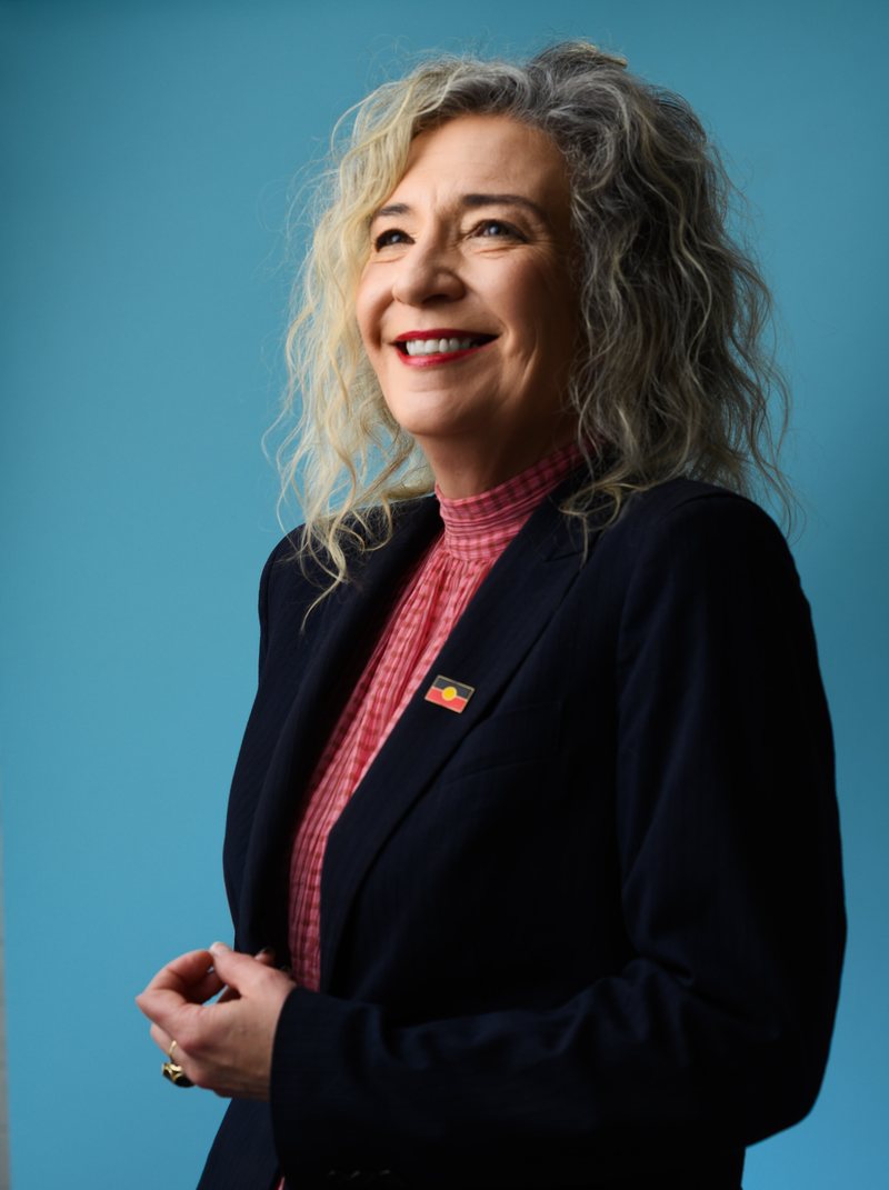 Melissa Lowe, smiling and looking off to one side, in a navy blazer over a pink checked blouse with an Aboriginal flag pin on the lapel, against a blue background.